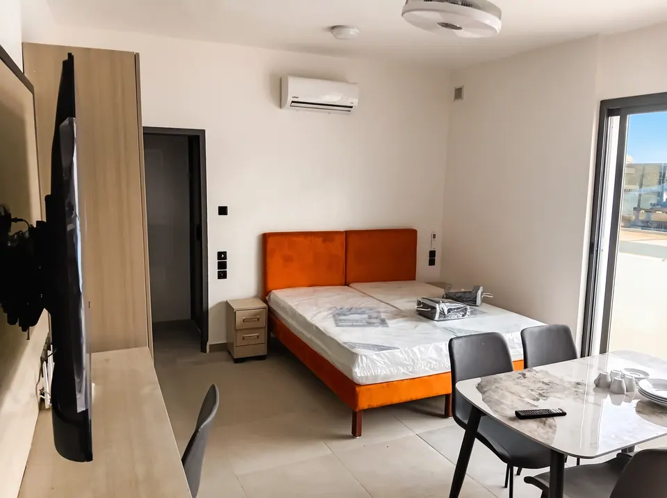 Chambre lumineuse avec un lit à tête de lit orange, un climatiseur mural, une table avec chaises et une porte-fenêtre