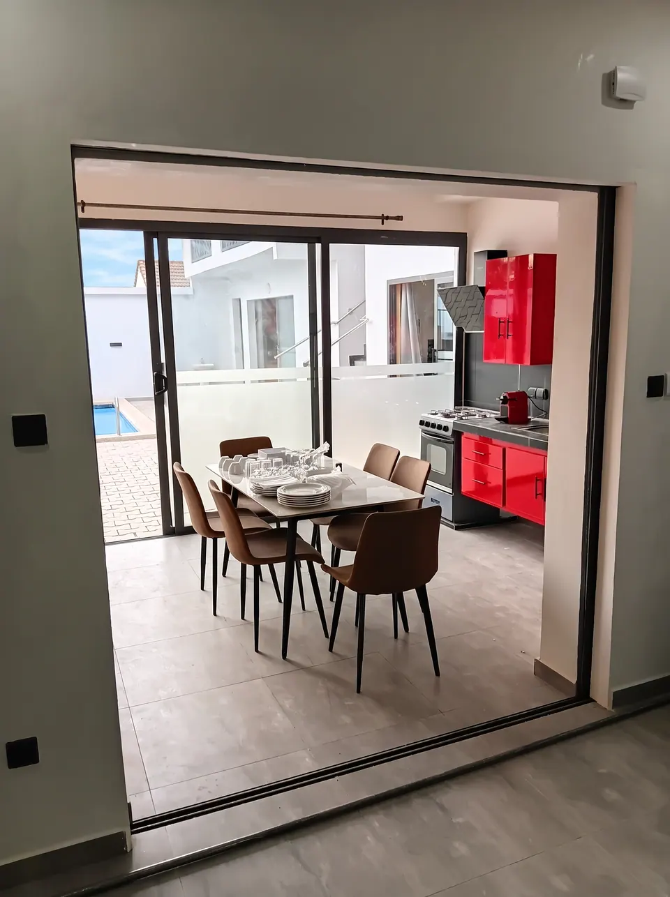 Table et chaises brunes devant un coin cuisine rouge, avec une baie vitrée donnant sur la cour